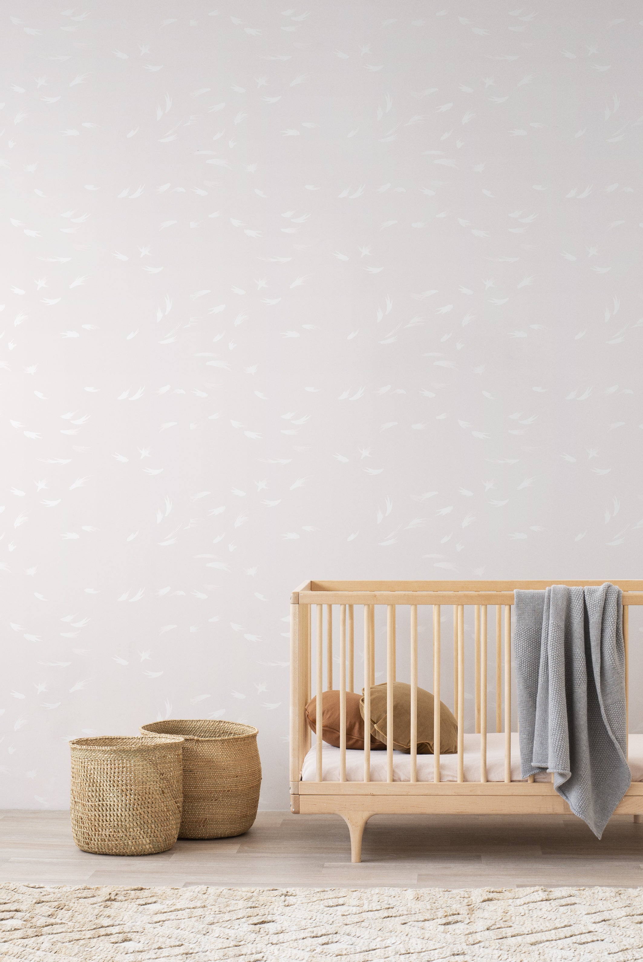 Falling Feather wallpaper by Emma Hayes, delicate white feathers on a warm blush-greige ground, styled in a nursery with timber cot and baskets