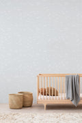 Falling Feather wallpaper by Emma Hayes, white feathers on a cool dove-grey ground styled in a nursery behind a timber cot with grey throw