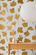 Petals Pressed wallpaper by Emma Hayes in mustard gold, bold painted petal silhouettes on pale grey, styled behind a timber