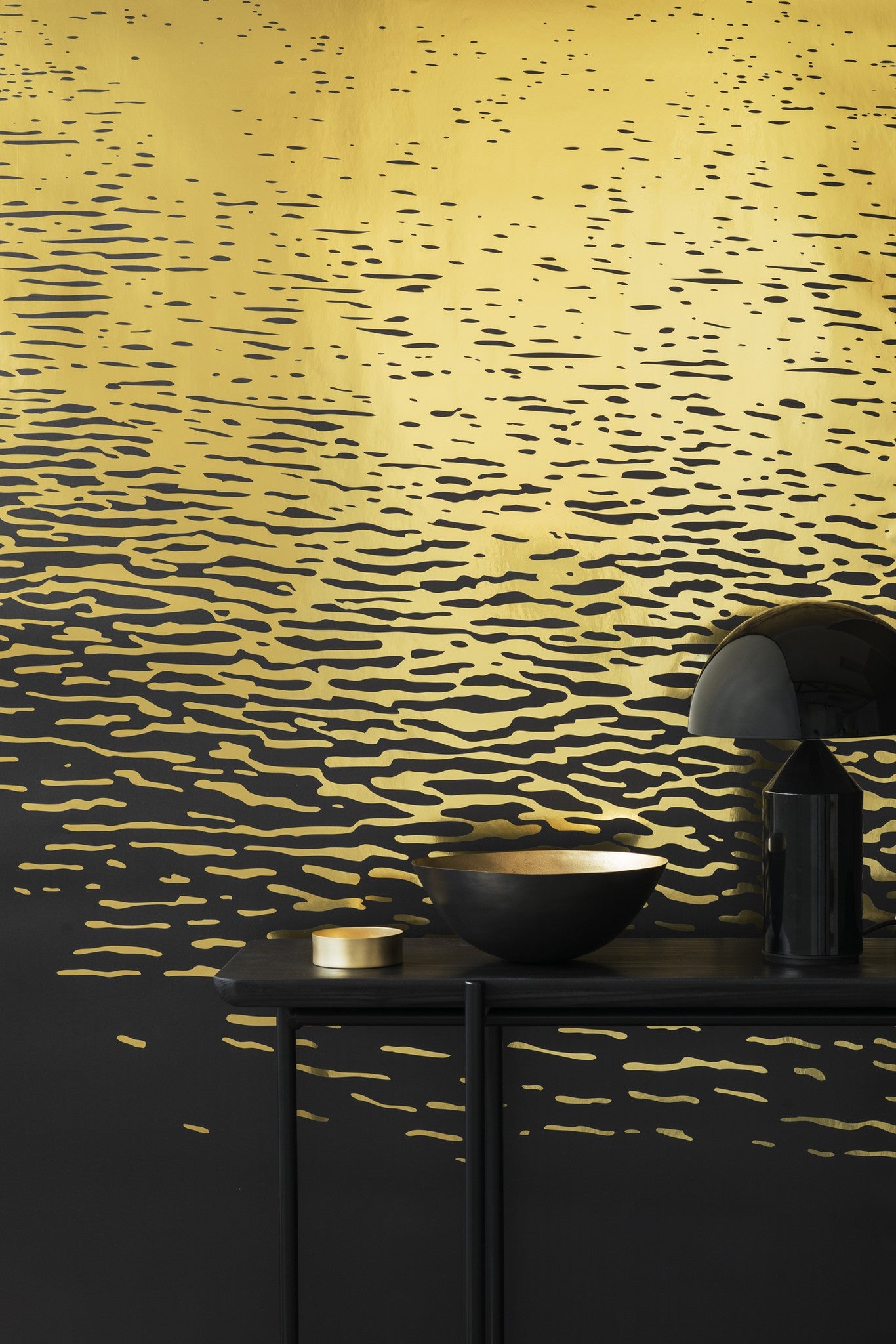 Reflection wallpaper by Emma Hayes in metallic gold on black, shimmering light-on-water ripples, styled behind a dark console with a brass bowl and lamp
