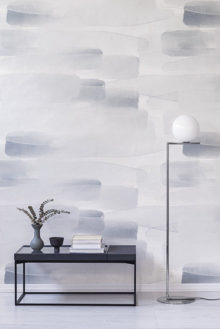 River wall mural by Emma Hayes in soft grey-blue watercolour brushstrokes, styled in a living room above a black console with vase and globe lamp