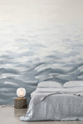 Seascape wall mural by Emma Hayes in Steel, soft grey rolling-wave brushstrokes across a bedroom feature wall behind a linen-dressed bed and lamp