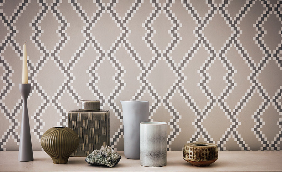 Danton wallpaper by Romo, a stepped ikat trellis in grey and stone, styled behind a candlestick and a group of ceramic