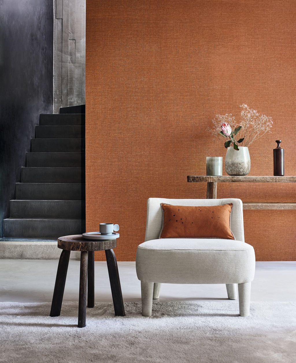 Carioca wallpaper by Casamance in burnt orange, a plain woven texture styled beside a black stone staircase with a cream boucle chair