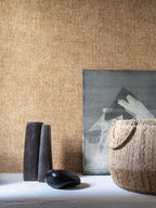 Diola wallpaper by Casamance in tan and gold, a plain woven texture styled behind dark sculptural vessels and a woven basket