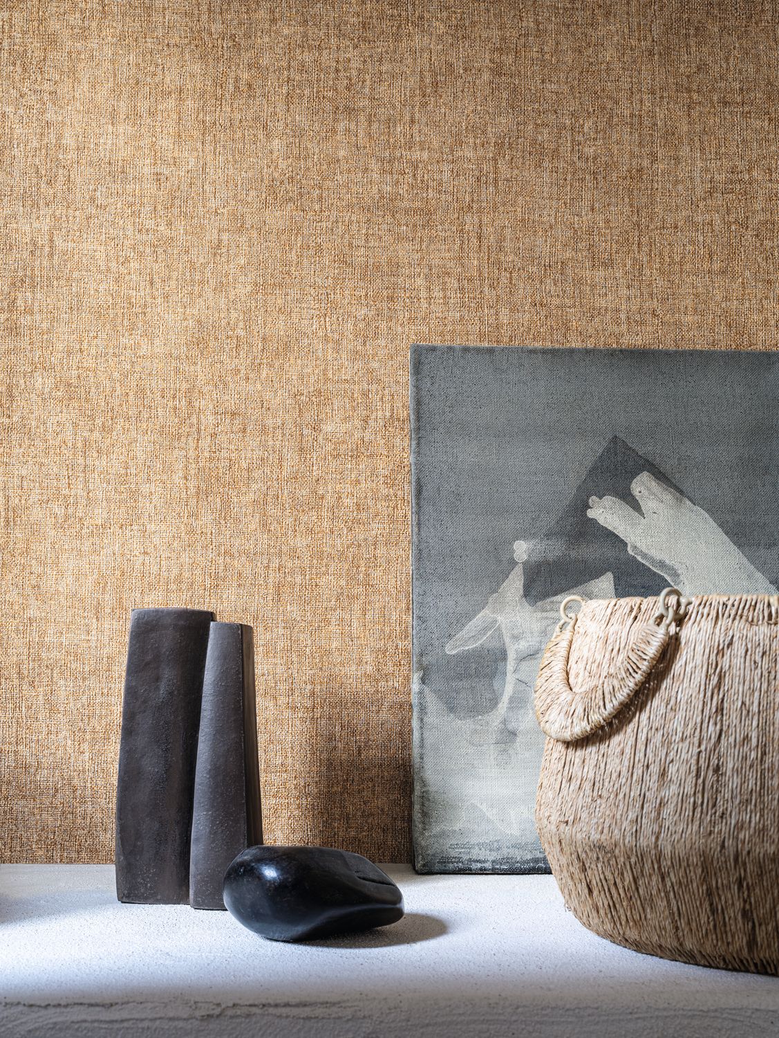 Diola wallpaper by Casamance in tan and gold, a plain woven texture styled behind dark sculptural vessels and a woven basket