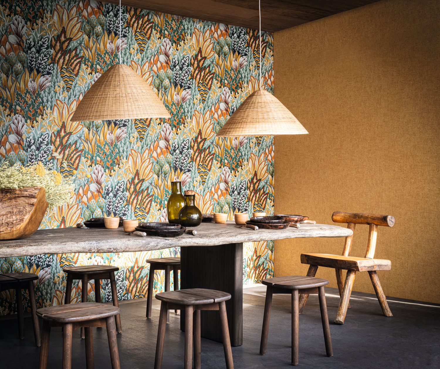 Avicennia wallpaper by Casamance in emerald and mustard, a dense protea and foliage print styled in a dining room with woven rattan pendants and a stone table