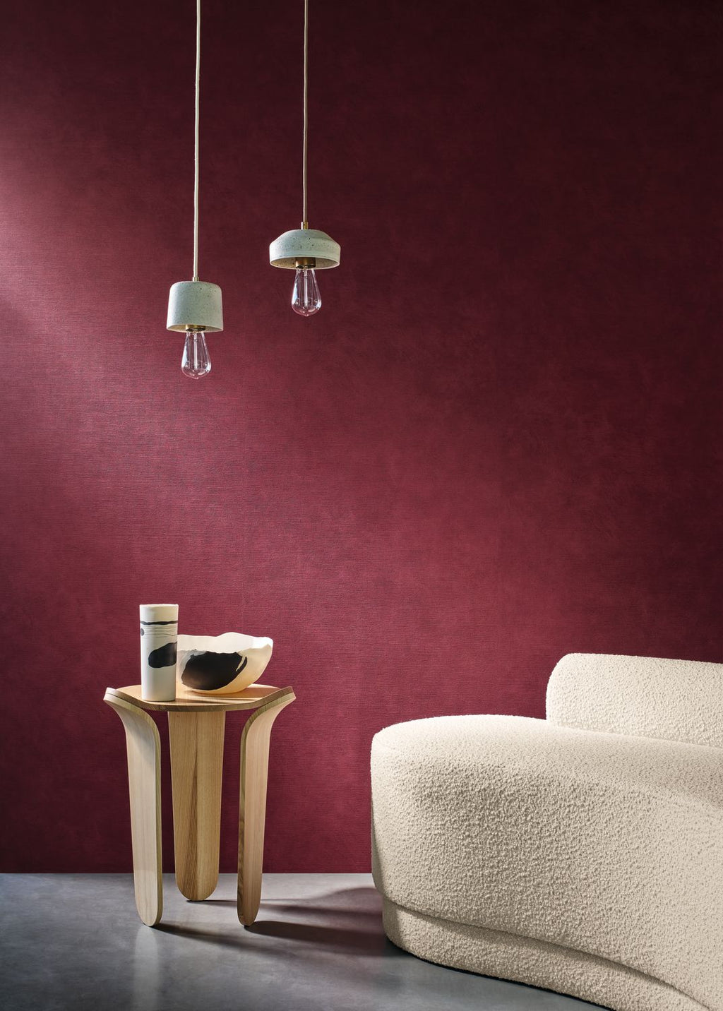 Argile wallpaper by Casamance in deep burgundy, a plain chalky texture styled behind a cream boucle sofa with glass pendant lights