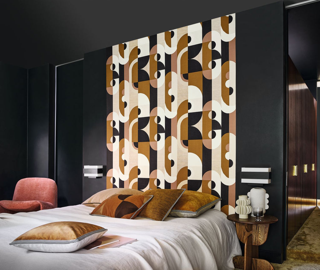 Vassily wallpaper by Casamance in bronze and ochre, a bold geometric design styled on a panel behind a bed in a dark room