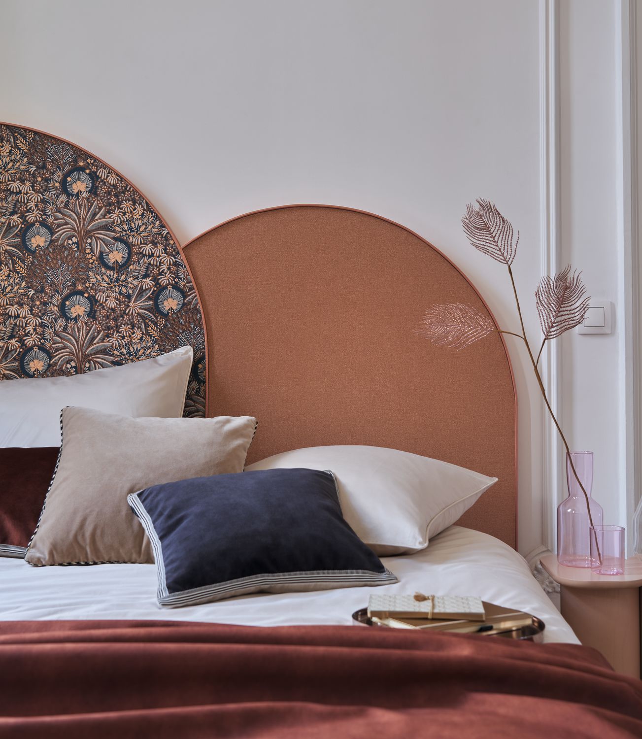 Octa wallpaper by Casamance in rust, a plain finish styled as an arched headboard panel above a bed with cream and navy cushions