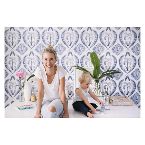 Blue Tropics wallpaper by Cass Deller — blue palm-tree damask styled behind a smiling mother and child in a bright living room