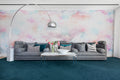 Coral Clouds pink mural by Rebel Walls styled in a lounge behind a long grey sofa on teal carpet